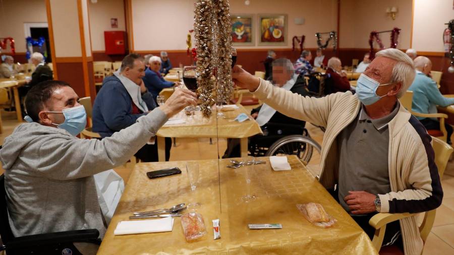 Dos residentes del centro Domus VI San Lázaro, en Santiago, brindan con una mampara en el medio que los separa durante la cena de Nochebuena. Foto: Lavandeira J.r.