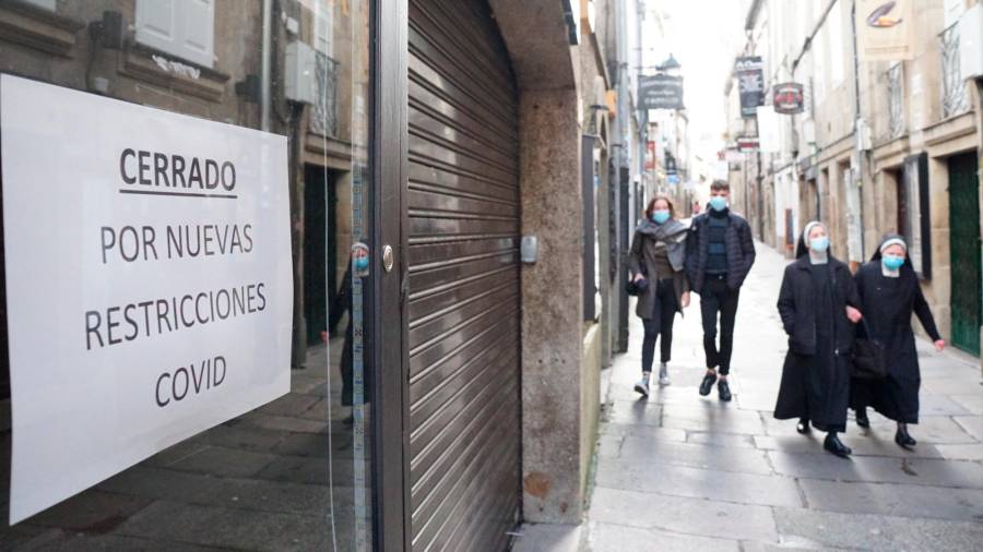 Paseantes ayer en una calle compostelana en la que un local comercial está cerrado por restricciones de la covid-19. Foto: Fernando Blanco